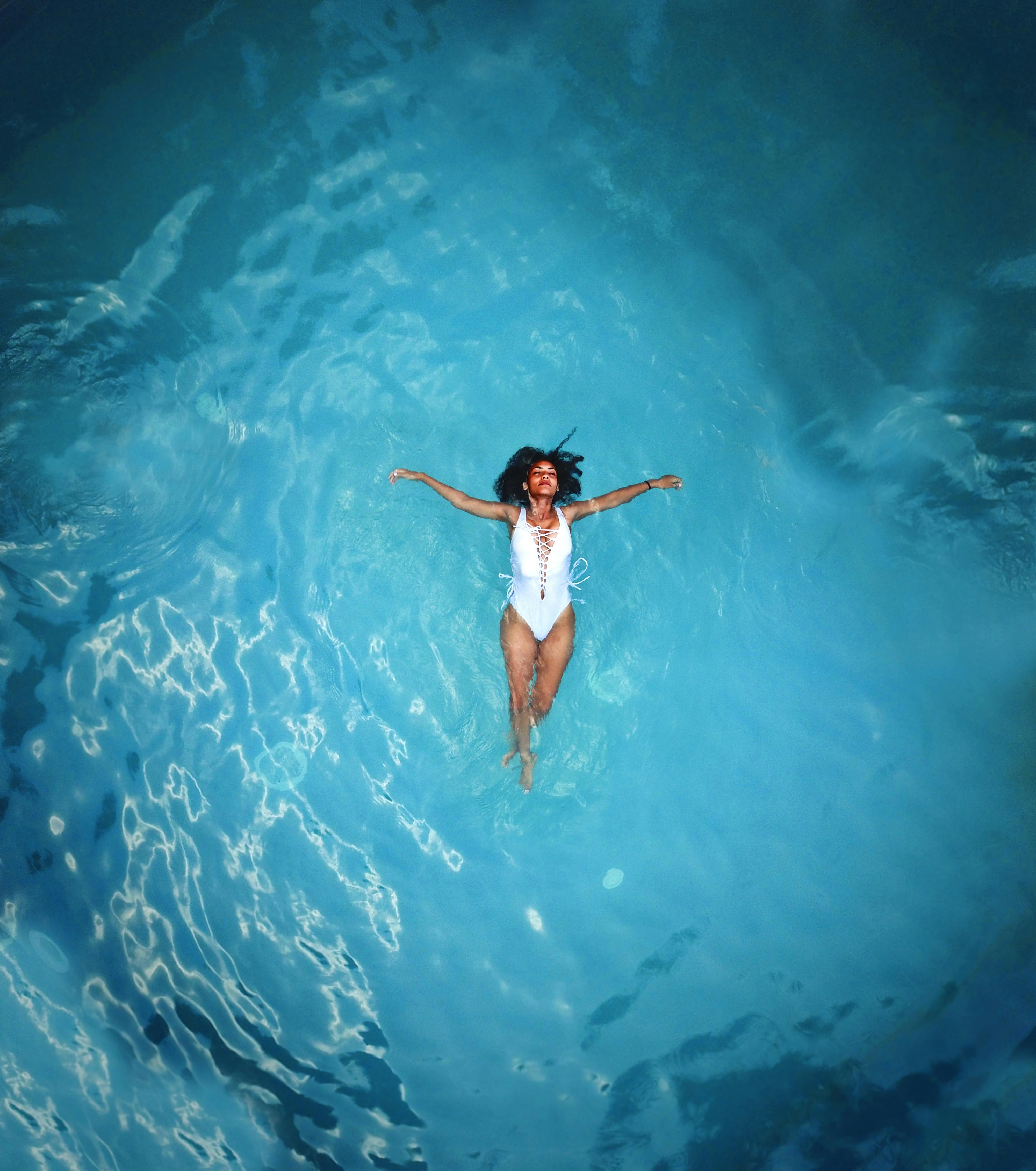 Woman in a white swimsuit floating peacefully on her back in a calm, clear blue pool, arms stretched out, with a serene and relaxed expression on her face.